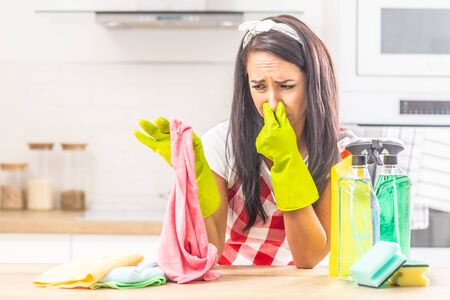 Female holding her nose with fingers upon smelly rag held in the other hand, leaning on kitchen desk, with cleaning stuff surrounding her.の写真素材