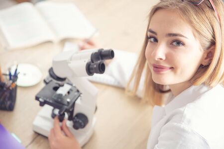 Beautiful young woman looking up to the camera while working with a microscope, writing down notes.の写真素材