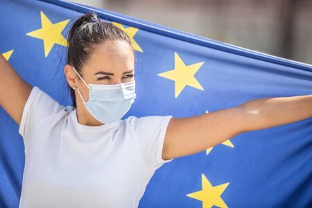 Female in face mask looking to the side hold a flag of the European union.
