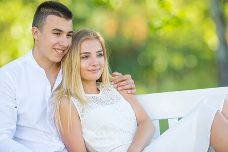 Happy young couple dressed in white looking the same direction on a park bench smiling. Boy holding the girl around her shoulder.の写真素材