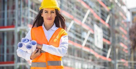 Female architect wearing orange safety vest and a yellow helmet holds rolled construction plans...