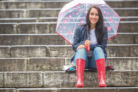 Pretty girl awaits her date sitting on stone stairs protecting herself from rain by an umbrella.の写真素材