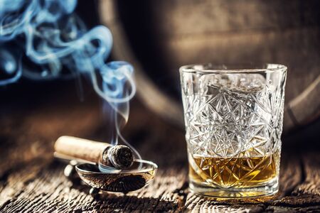 Whisky with no ice in a cup with burning cigar and an old wooden barrel in the background.の写真素材