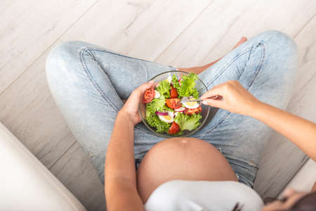 Top view of a pregnant woman sitting with a fresh salad bowl in her lap.