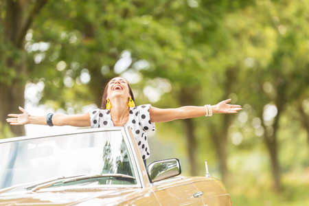Happy couple on a summer roadtrip with girl sitting on top of a convertible back seat, arms open,...