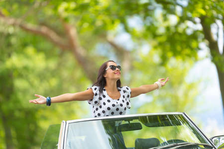 Woman in sunglasses sits on top of the convertible, arms wide open, soaking up the sun and fresh...