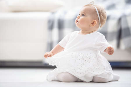 Small girl dressed in white sits on a floor at home, looking up.