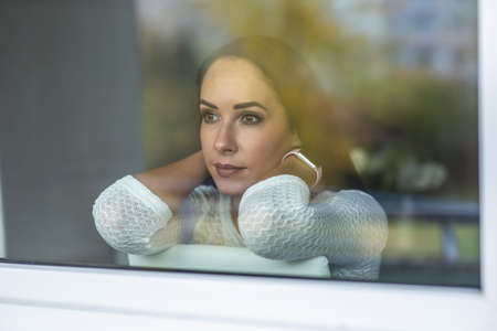 Isolation sadness by a young female sitting behind the window in the apartment, looking out.
