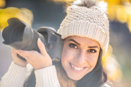 Smiling good-looking female photographer in a pompom hat takes pictures outside on a sunny fall day.