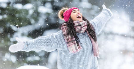 Catching snowflakes to open mouth outdoors by a happy woman in sweater, scarfs and a winter hat.