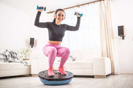 Fit beautiful woman in a training outfit and headphones squats at home on a balance ball holding...