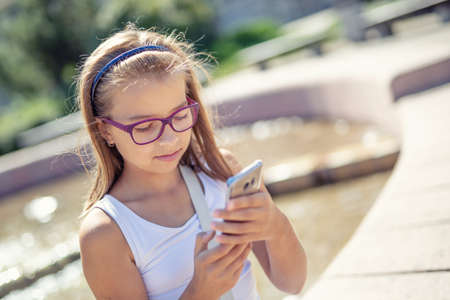 Young pre-teen blonde girl looks into the mobile phone in her hand sitting on the edge of a fountain...