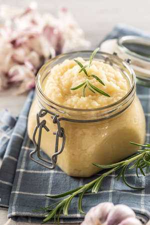 Aromatic garlic paste in a glass jar laid on rustic kitchen cloth with bulbs and peeled cloves and rosemary.の写真素材