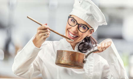Smiling female chef in professional outfit tastes food from wooden spoon holding a copper pot.の写真素材