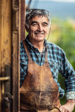 Older and experienced craftsman stands smiling in front of the workshop in checkered shirt and leather apron.の写真素材