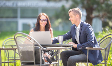 Office male and female colleagues discuss work over an open laptop and a takeaway coffee cup outdoors.の写真素材
