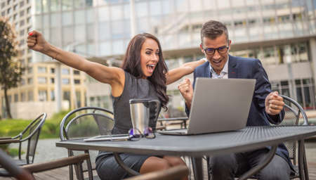 Male and female coworkers showing thrilled enthusiasm over successful deal just done on a laptop in fron of them outdoors in the city.の写真素材