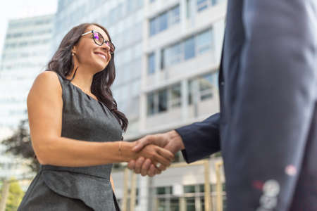 Two people in business clothes shake hands surrounded by office buildings.の写真素材