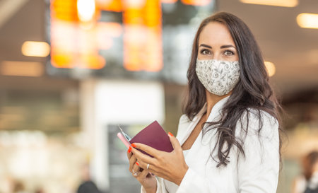 Brunette with face mask checks her passport before travelling abroad with an airport information panel behind her.の写真素材