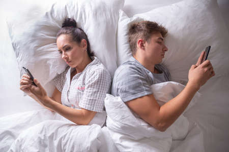 Couple separated in bed by digital technology as they both lie backs to each other looking at their cell phones.の写真素材