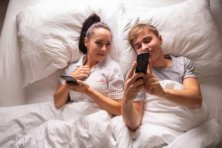 Top view of a couple sharing interesting and funny things on their mobile phones while lying in white bedding before sleep.の写真素材