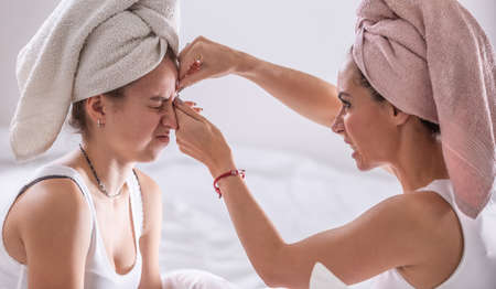 Mom squeezes out acne from her daughter's face, both wearing towels on their heads.の写真素材