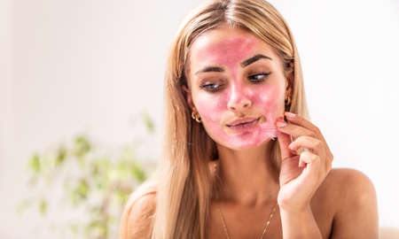 Caucasian blonde girl applies pink peeling mask on her face indoors.