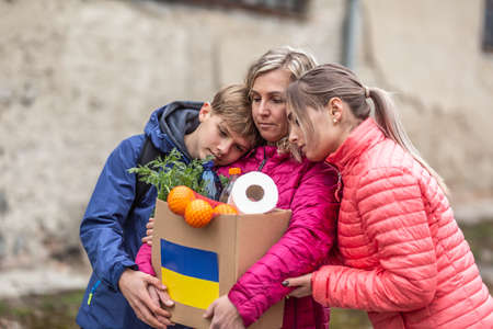 Two teenage kids comfort sad mother in Ukraine holding box full of humanitarian aid from Europe.