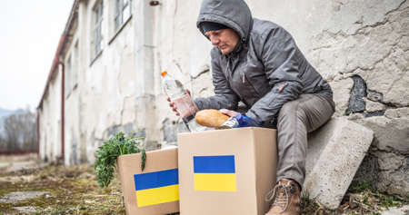 Older man in warm clothes sits on the edge of derilicted building taking bread and water out of humanitarian aid package for Ukraine.の写真素材