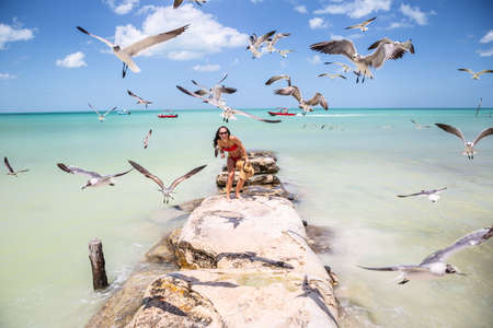 Woman in swimsuit stands on rocks next to the exotic sea laughing as flock of seagulls fly closely...