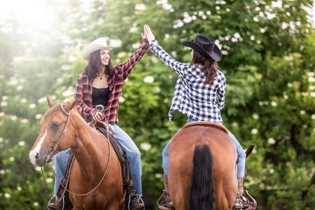 Two cowgirl friends with horses facing the opposite direction give a high five.の写真素材