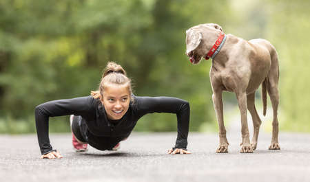 Young woman doing pushups outdoors smiles at her dog observing her.の写真素材