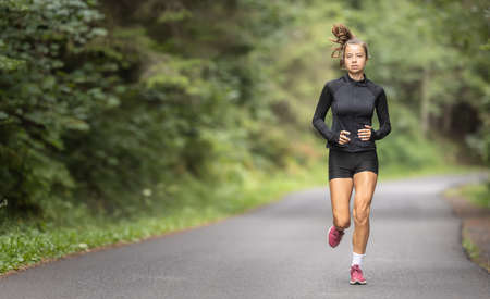 Fit young girl in black sportswear and red shoes runs on an empty asphalt road in the nature.の写真素材
