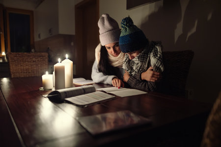 Woman and her son sit at the tabel at home dressed in winter caps and shawls shivering from cold due...