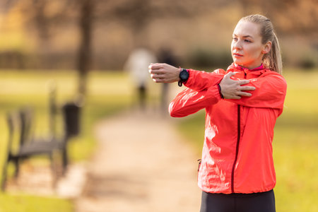Young woman warming up before jogging, doing upper body stretching in the park.