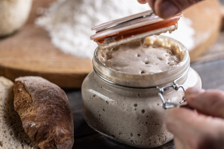 Male hands open a jar with active yeast, flour and fresh bread and pastries in the background.の写真素材