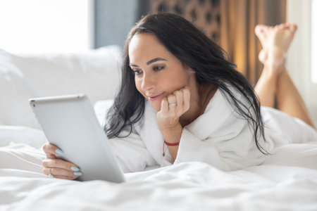 A good-looking brunette is lying on the bed in a bathrobe and using a tablet for a dating site.の写真素材