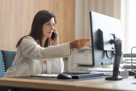 Executive female manager in a suit works at a computer in the office and gives tasks to her teamの写真素材
