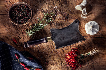 An old butchers cleaver on a cutting board surrounded by chili peppers garlic and rosemary - Top of view.の写真素材