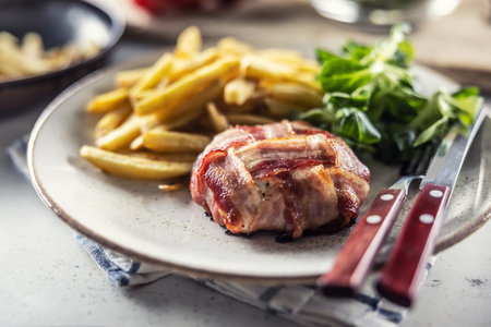 Bacon wrapped meat served with french fries and salad on a plate with cutlery.の写真素材
