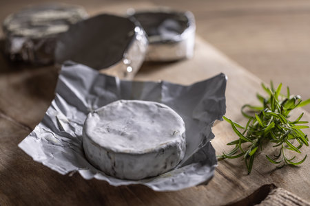 Packages of camembert cheese with one of them open next to rosemary on a wooden board.の写真素材