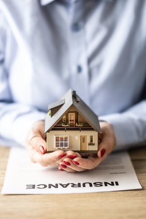House insurance concept displayed via female hands holding a model of a building on top of an insurance agreement.の写真素材