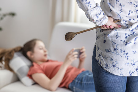 Adolescent girl ignores her mother standing upset with a wooden spoon next to her and keeps being on her mobile phone.の写真素材