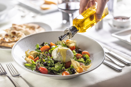 A hand of a person pours olive oil on top of Buffalo mozzarella ball on top of a healthy Italian salad.の写真素材