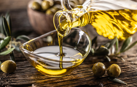 Pouring olive oil from carafe to the glass bowl and in background are olive branches put on table.の写真素材