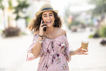 A smiling and happy young woman is walking through the city, talking on the phone and holding a takeaway coffee in her hand.の写真素材