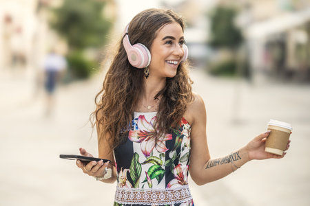 Young beautiful smiling female tourist is using headphones listening to music and holding takeaway coffee in her hand.の写真素材