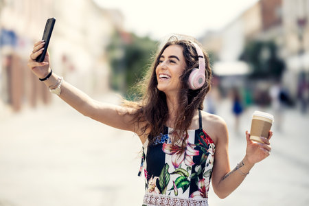 Young beautiful female tourist is taking a selfie, using headphones, listening to music and holding a takeaway coffee in her hand.の写真素材