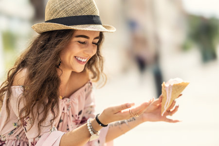 Beautiful young lady has melted ice cream on her fingers, sitting on a bench during a hot summer day.の写真素材