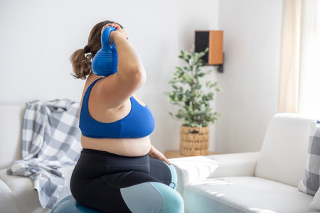 Overweight lady doing sports exercise in living room. She is sitting on a fitness ball and holding a dumbbell on her shoulder.の写真素材